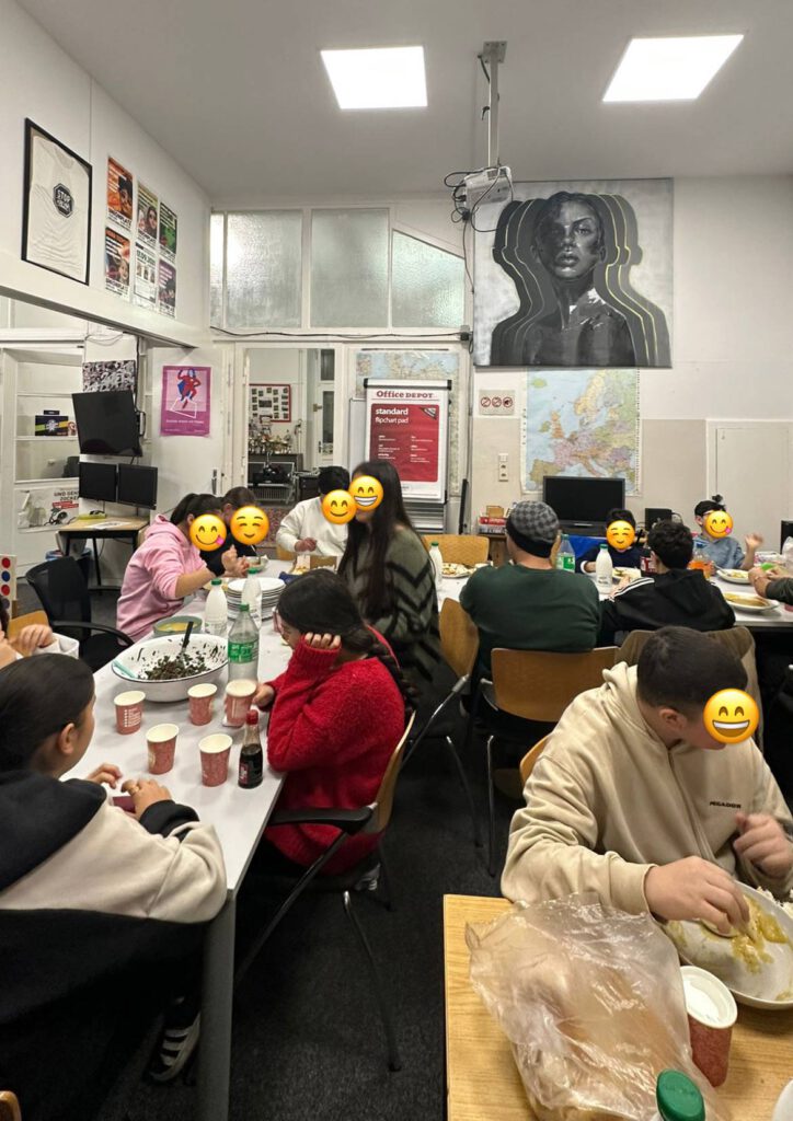 Innenraum eines Raumes mit mehreren Menschen, die an Tischen sitzen und gemeinsam essen. Die Gesichter der Personen sind mit Smileys bedeckt. Es sind verschiedene Getränke in Flaschen und Bechern auf den Tischen. An der Wand hängen Poster, ein Weltkarte und ein großes Gemälde einer Frau. Im Hintergrund sind Computer und Büromaterial sichtbar, was auf eine Gemeinschafts- oder Arbeitsraum hinweist.