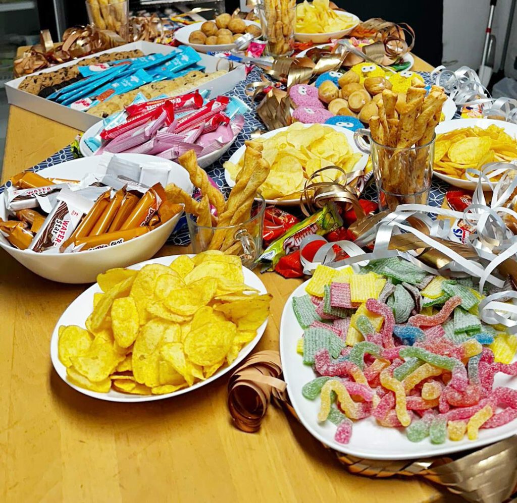 Tisch mit einer Vielzahl von Snacks und Süßigkeiten, darunter Chips, Gummibärchen, Croissants und Kekse, alles in weißen Schalen und Tellern angerichtet. Im Hintergrund sind weitere Leckereien und Dekorationen sichtbar.