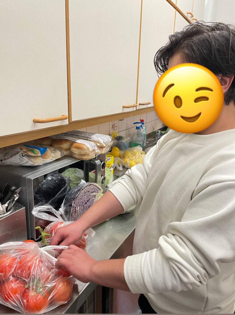 Person steht in einer Küche vor einer Arbeitsfläche, auf der mehrere Einkaufstüten mit frischem Gemüse und Obst liegen. Die Person greift nach einer Tomate aus einer transparenten Plastiktüte. Im Hintergrund sind Küchenutensilien, ein Mikrowellengerät und eine Wasserflasche sichtbar.