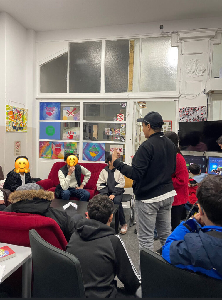 Gruppen von Menschen sitzen in einem hellen Raum und hören einem Sprecher zu, der vor ihnen steht. Die Gesichter der Personen sind mit Smileys bedeckt. Der Raum ist dekoriert mit bunten Kunstwerken an den Wänden, und im Hintergrund sind Fenster und eine Tür zu sehen.