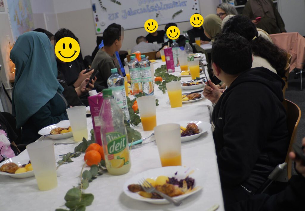 Das Bild zeigt eine Gruppe von Menschen, die an einem langen Esstisch sitzen. Der Tisch ist mit einer weißen Tischdecke bedeckt und ist festlich geschmückt mit einer Kette aus grünen Blättern und Orangen. Auf dem Tisch stehen mehrere Flaschen Wasser, Orangensaft und andere Getränke. Es gibt Teller mit Essen, darunter Hähnchen, Kartoffeln und Rotkohl. Die Menschen essen, trinken und unterhalten sich. Im Hintergrund sind weitere Personen, einige tragen Brillen und Kopfbedeckungen. An der Wand hängt eine Tafel mit der Aufschrift "zur Jahresabschlussfeier". Die Gesichter der Menschen sind durch Smileys ersetzt. Die Atmosphäre wirkt fröhlich und gesellig.