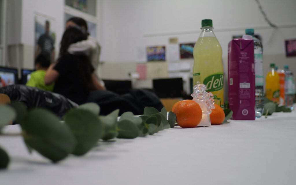 Das Bild zeigt eine Tischdeko mit mehreren Gegenständen. Im Vordergrund liegen grüne Blätter, zwei Orangen, ein kleines transparentes Schneekugel- Dekor und andere Früchte. Auf dem Tisch stehen Getränke, darunter eine grüne Flasche mit einem grünen Deckel, eine pinkfarbene Verpackung, sowie eine Wasserflasche. Im Hintergrund sind Menschen, die sich unterhalten, und es gibt einen Raum mit Bildern an der Wand. Die Szene wirkt wie eine Zusammenkunft oder Feier in einem Innenraum.