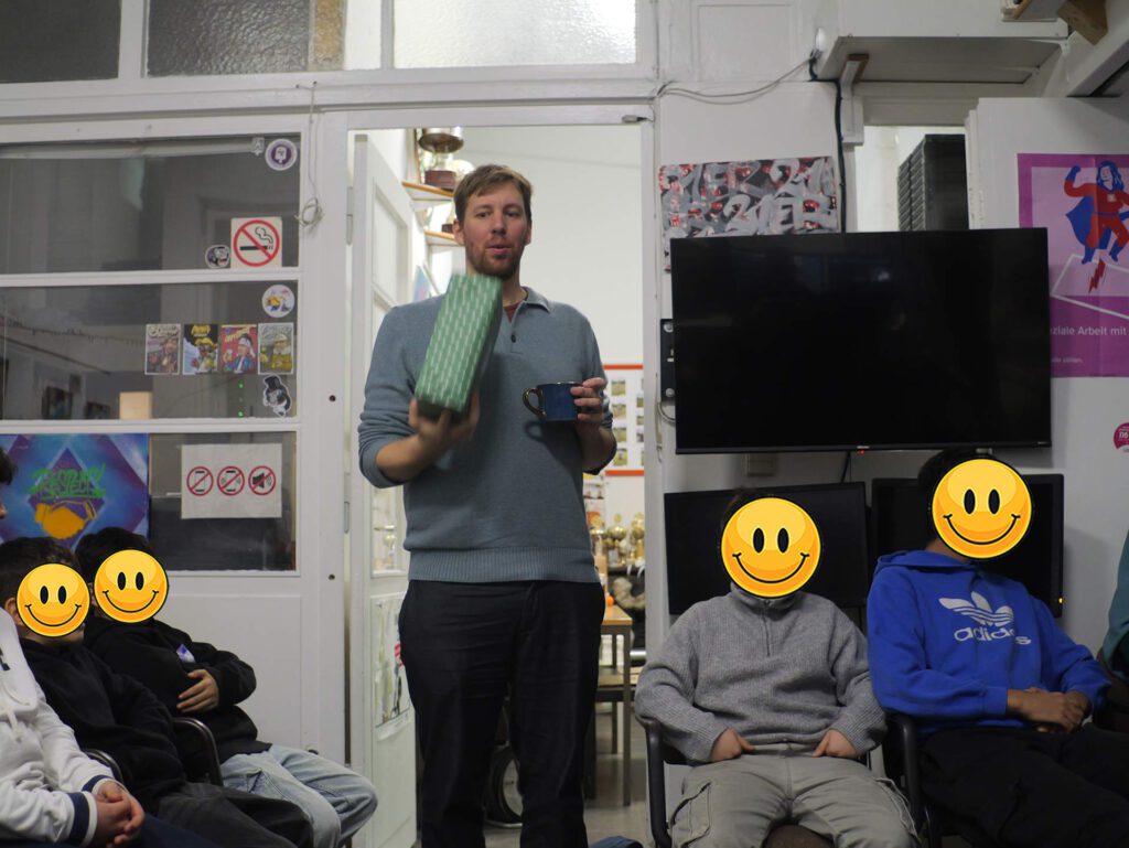 Bild zeigt einen Mann, der vor einer Gruppe von Menschen steht. Er hält ein Geschenk in der Hand und eine Tasse. Die Kinder sitzen in Stühlen und hören ihm zu. Auch ihre Gesichter sind durch lachende Smiley-Gesichter ersetzt. Im Hintergrund sieht man eine Tür mit Fenstern, an denen Aufkleber und Bilder kleben. Es sieht aus wie eine kleine Feier oder ein Treffen in einem Raum.