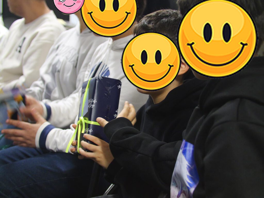 Bild zeigt eine Gruppe von Kindern, die in einer Reihe sitzen. Die Gesichter der Kinder sind durch große, lachende Smiley-Gesichter ersetzt. Einige Kinder halten Geschenke in den Händen. Sie tragen unterschiedliche Kleidung, zum Beispiel eine weiße Jacke, eine schwarze Jacke und eine graue Jacke. Es sieht aus, als ob die Kinder bei einer Feier oder einem Fest sind.