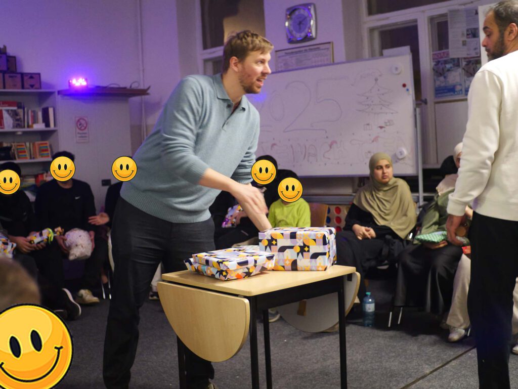 Bild zeigt eine Person, die vor einer weißen Tafel steht und einen Marker in der Hand hält. Die Tafel hat das Wort „Wichtel“ geschrieben. Im Hintergrund sitzen mehrere Menschen, die sich angucken. Die Gesichter der Personen sind durch Smileys ersetzt. Es sieht nach einer Aktivität oder einem Vortrag aus.