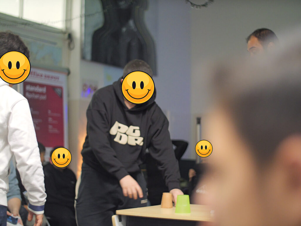 Bild zeigt eine Gruppe von Menschen, die in einem Raum stehen. Die Gesichter der Personen sind durch große, lachende Smiley-Gesichter ersetzt. Eine Person in der Mitte trägt ein schwarzes Hoodie mit weißen Buchstaben. Es sieht aus, als ob sie an einem Spiel oder einer Aktivität teilnehmen.