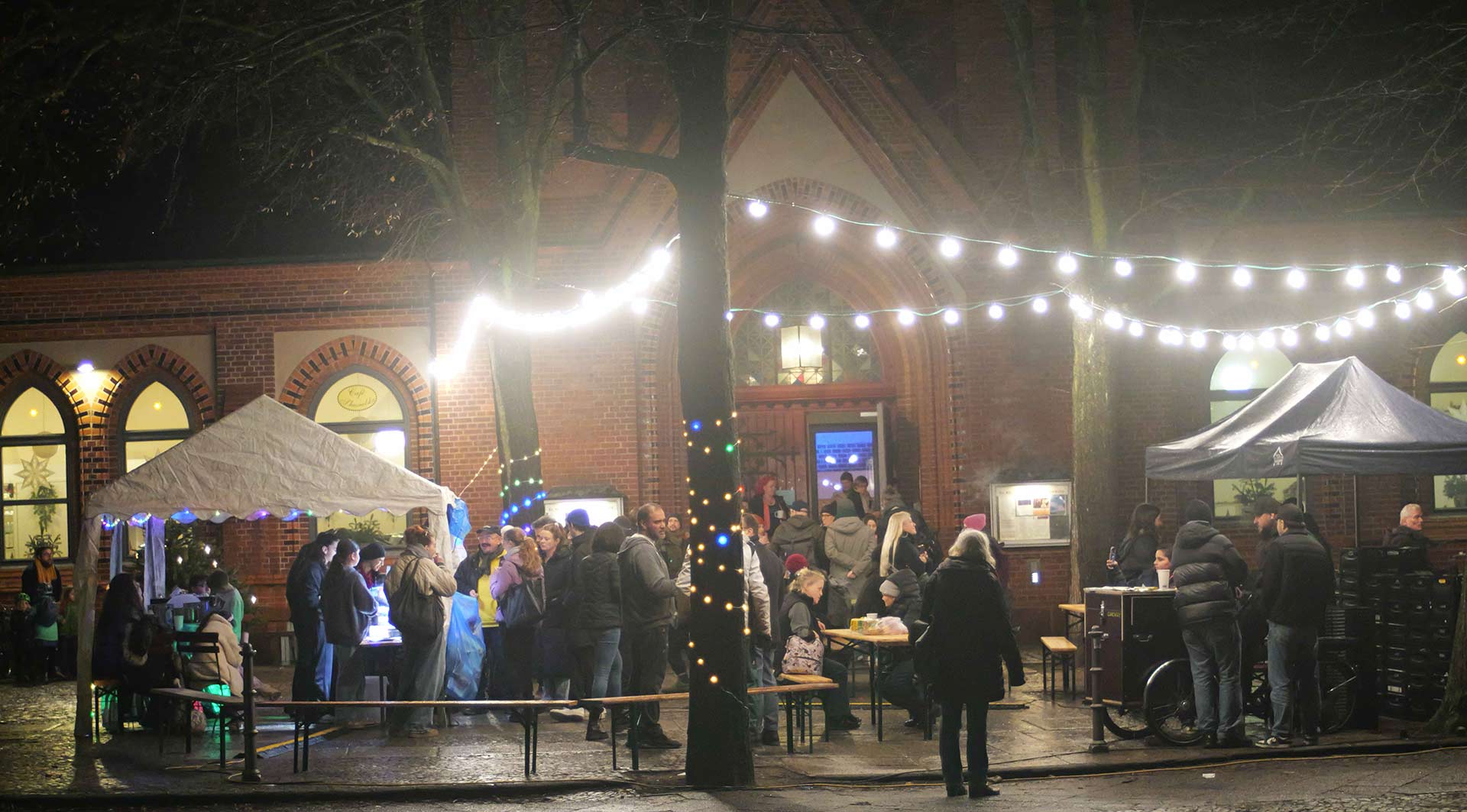 Auf dem Bild ist eine Menschenmenge vor einem roten Backsteingebäude zu sehen, das vermutlich eine Kirche oder ein historisches Gebäude ist. Es ist dunkel und es ist Nacht. Das Gebäude ist mit bunten Lichterketten dekoriert. Im Vordergrund stehen mehrere Tische und Bänke, einige Menschen sitzen, andere stehen in einer Warteschlange. Zwei Zelte sind aufgebaut, eines auf der linken Seite und eines auf der rechten Seite. Es gibt mehrere Personen, die an einem Stand im rechten Zelt bedient werden, darunter auch eine Person im Rollstuhl. Die Szene wirkt festlich, vermutlich handelt es sich um einen Weihnachtsmarkt oder ein ähnliches Fest. Es sind viele Menschen unterschiedlicher Altersgruppen zu sehen, die sich unterhalten und das Fest genießen.