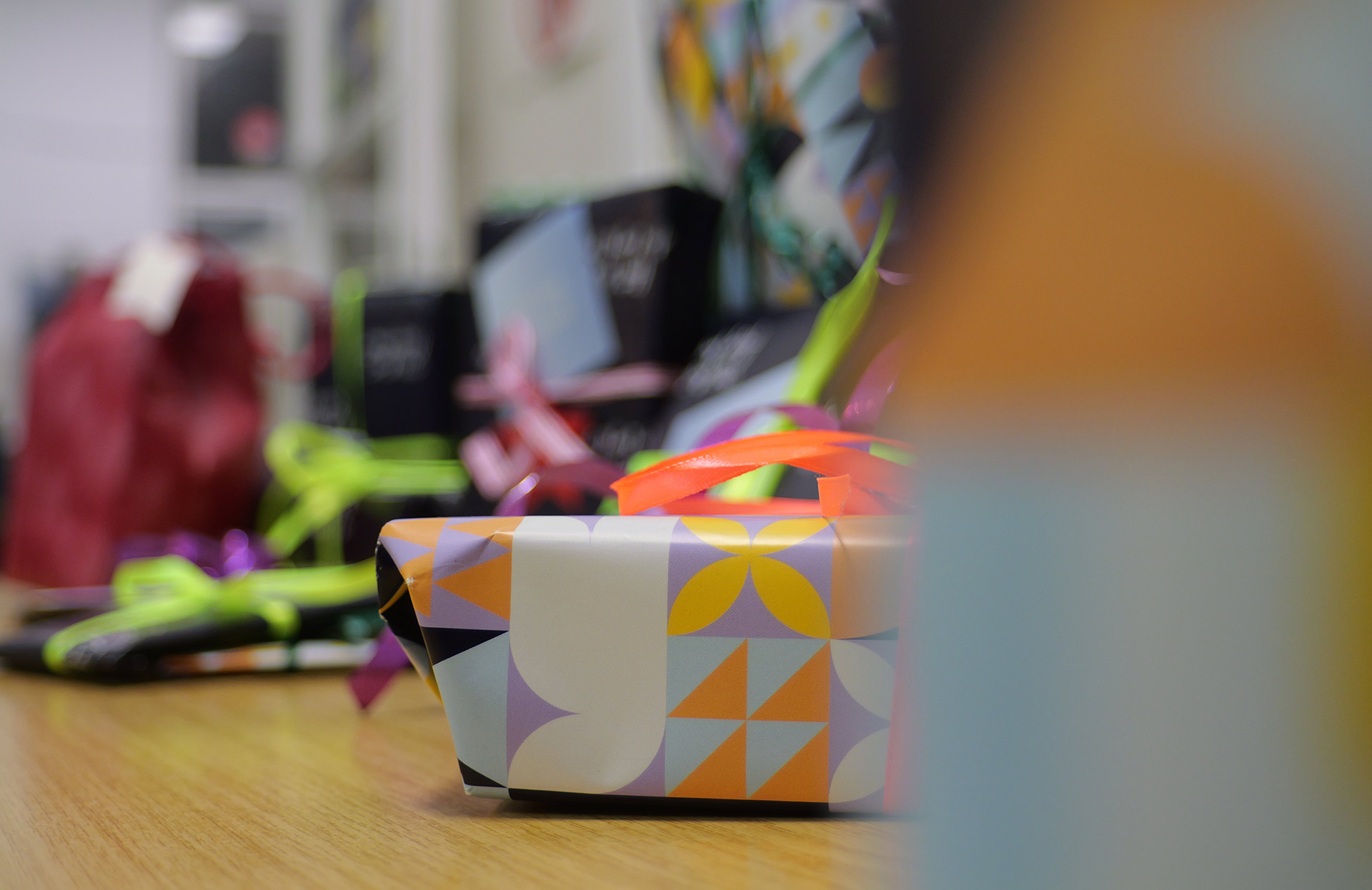 Das Bild zeigt eine Gruppe von Geschenken, die auf einem Tisch liegen. Im Vordergrund ist ein Geschenk mit buntem, geometrischem Muster in den Farben Orange, Lila, Weiß, Schwarz und Hellblau. Das Geschenk hat eine orangefarbene Schleife. Im Hintergrund sind weitere Geschenke, die mit bunten Schleifen und Bändern verziert sind. Die Geschenke sind unterschiedlich groß und in verschiedenen Farben eingepackt. Die Umgebung wirkt wie eine festliche Szene, möglicherweise für eine Feier oder einen besonderen Anlass.