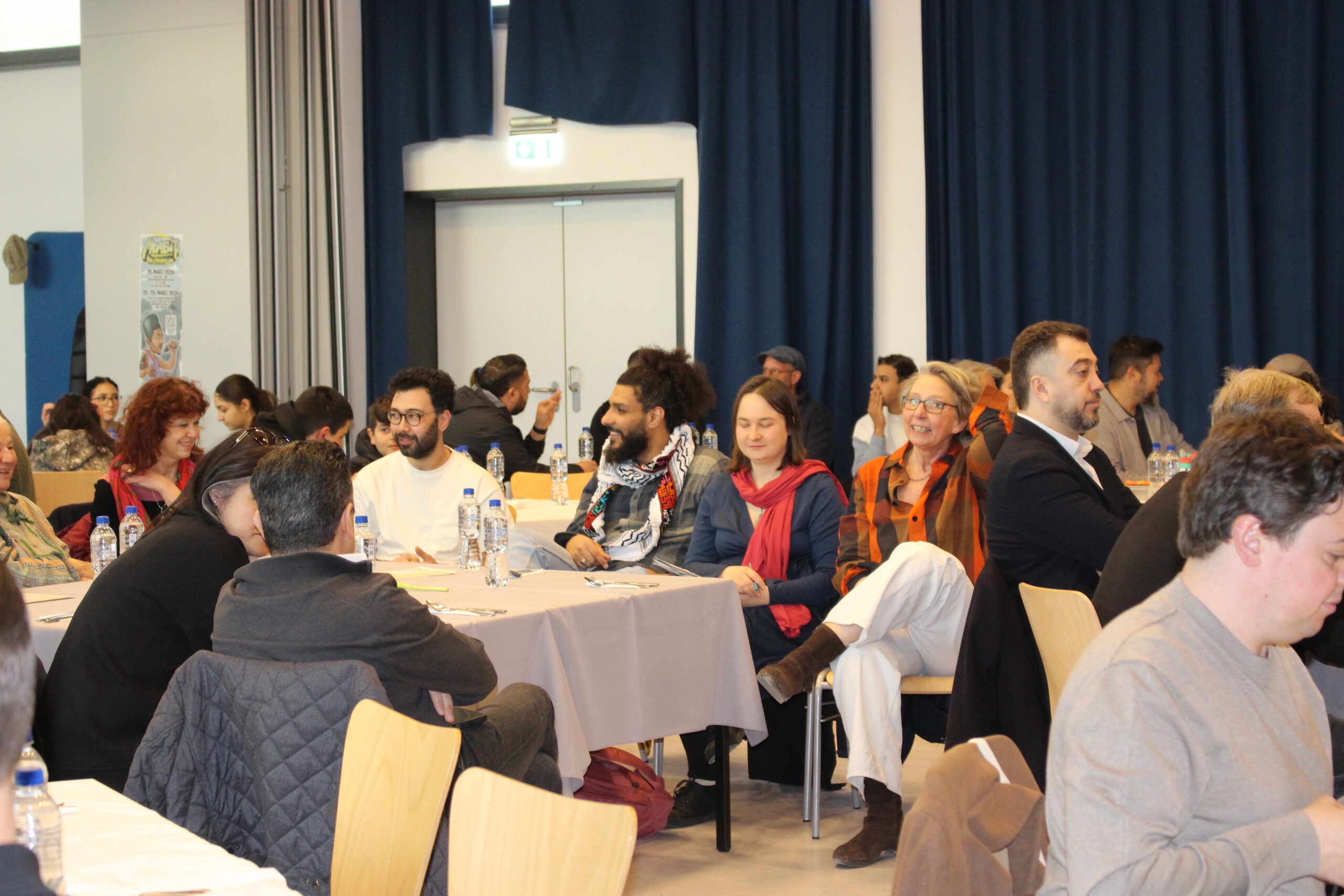 Ein Gruppenfoto von mehreren Menschen, die bei einer Veranstaltung an Tischen sitzen, sich unterhalten und lachen. Im Hintergrund sind dunkle Vorhänge und eine Tür sichtbar.